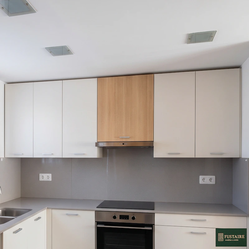 Fotografía de un diseño de cocina a medida moderna instalada en Barcelona por la carpintería Fustaire. Presenta frentes de armarios en blanco roto, una campana extractora con panel de madera clara, encimera gris, fregadero integrado y electrodomésticos en acero inoxidable, en un espacio funcional y luminoso